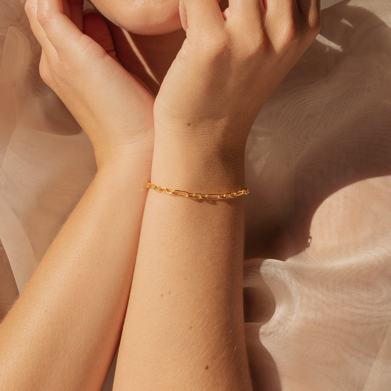A minimal jewelry look: the Roxy Paperclip Chain Bracelet adorns a wrist as hands gently touch their face, set against soft beige fabric.