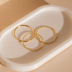 The Skinny Stacking Rings Set features three gold minimalist rings, including two with twisted designs, displayed on an oval white tray—perfect for a dainty, stacked look.