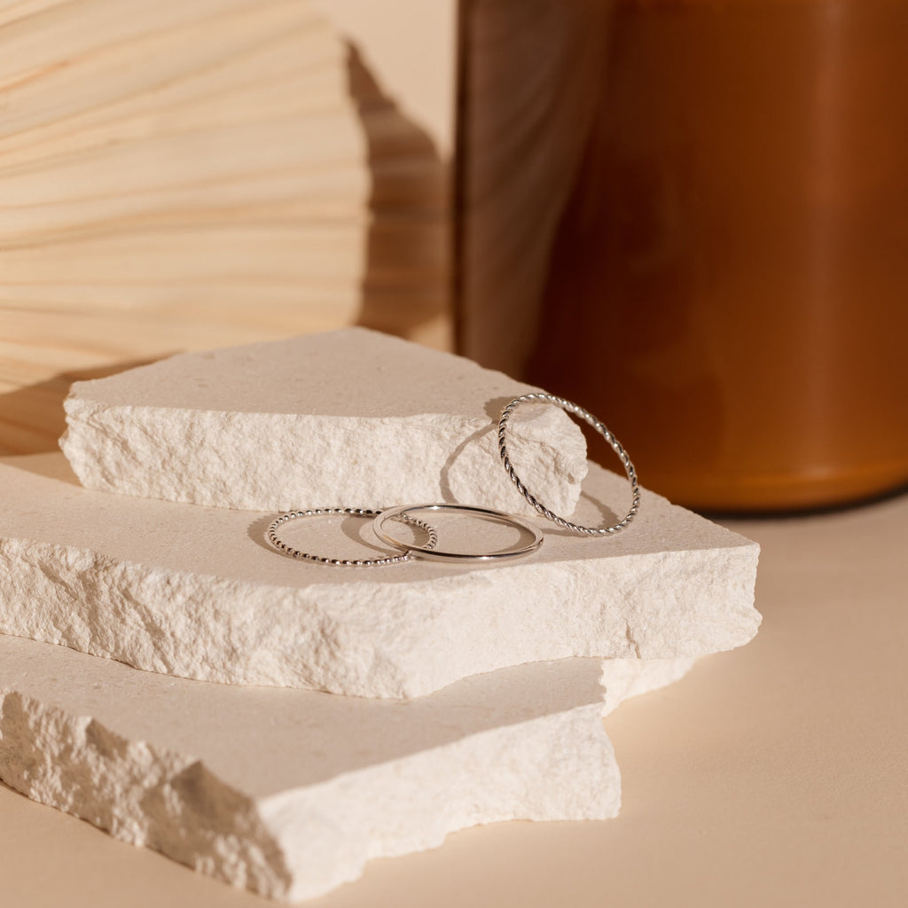 The Skinny Stacking Rings Set is displayed on white stone slabs, with a wooden object and brown vase in the background—perfect for those who love minimalist rings.