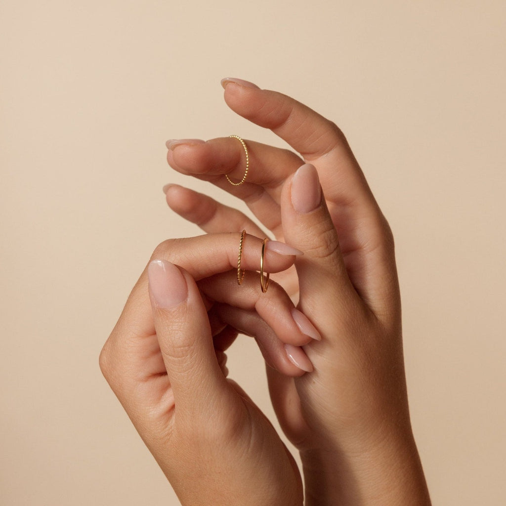 Two hands with nude nails wear the Skinny Stacking Rings Set and dainty gold bands, posed gracefully against a beige background.
