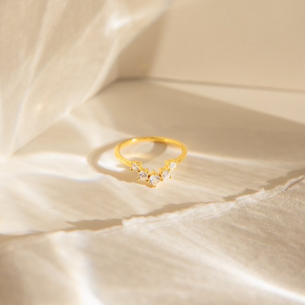 The Marina Chevron Ring, featuring a dainty gold band with small diamonds, is displayed on a softly lit white fabric background.