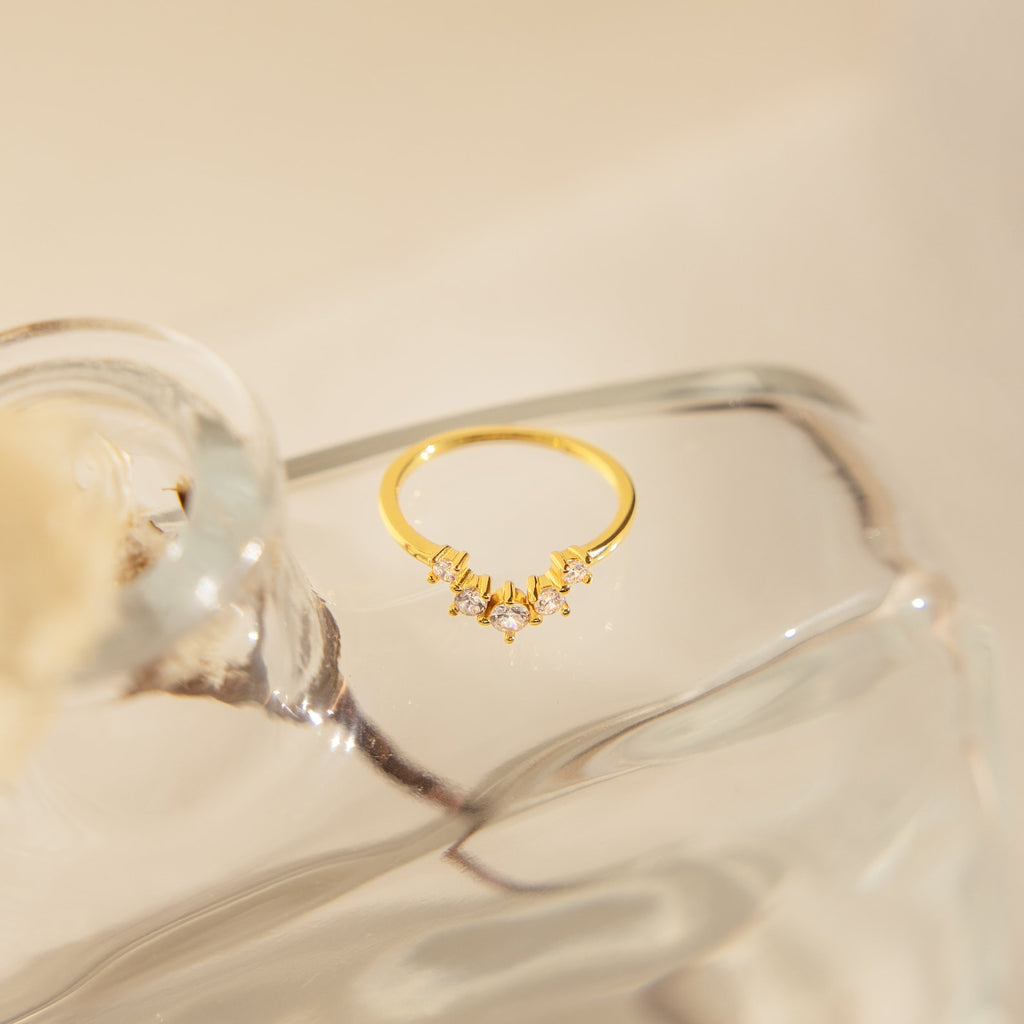 The Marina Chevron Ring, a delicate gold band adorned with shimmering stones, rests on a clear sculpted glass surface under soft lighting.