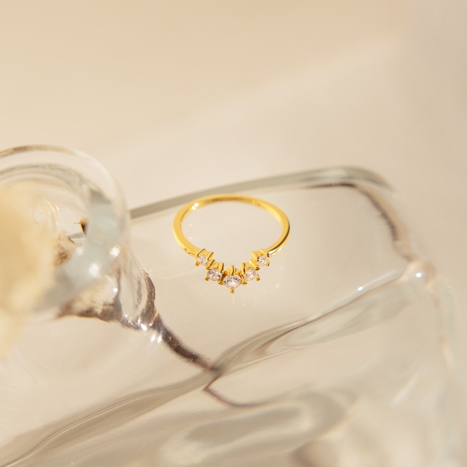 The Marina Chevron Ring, a delicate gold band adorned with shimmering stones, rests on a clear sculpted glass surface under soft lighting.