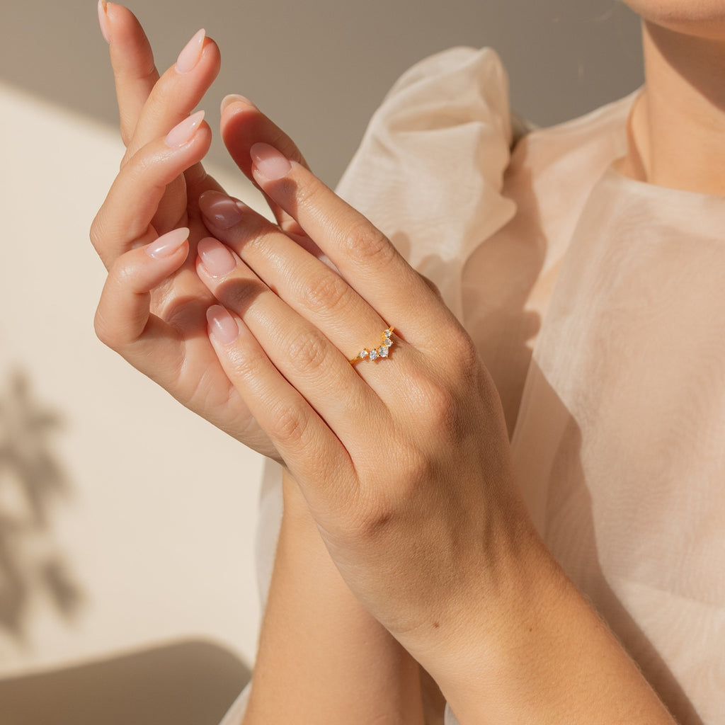 A woman in a sheer white blouse showcases the Marina Chevron Ring, a dainty band adorned with small gemstones on her finger.