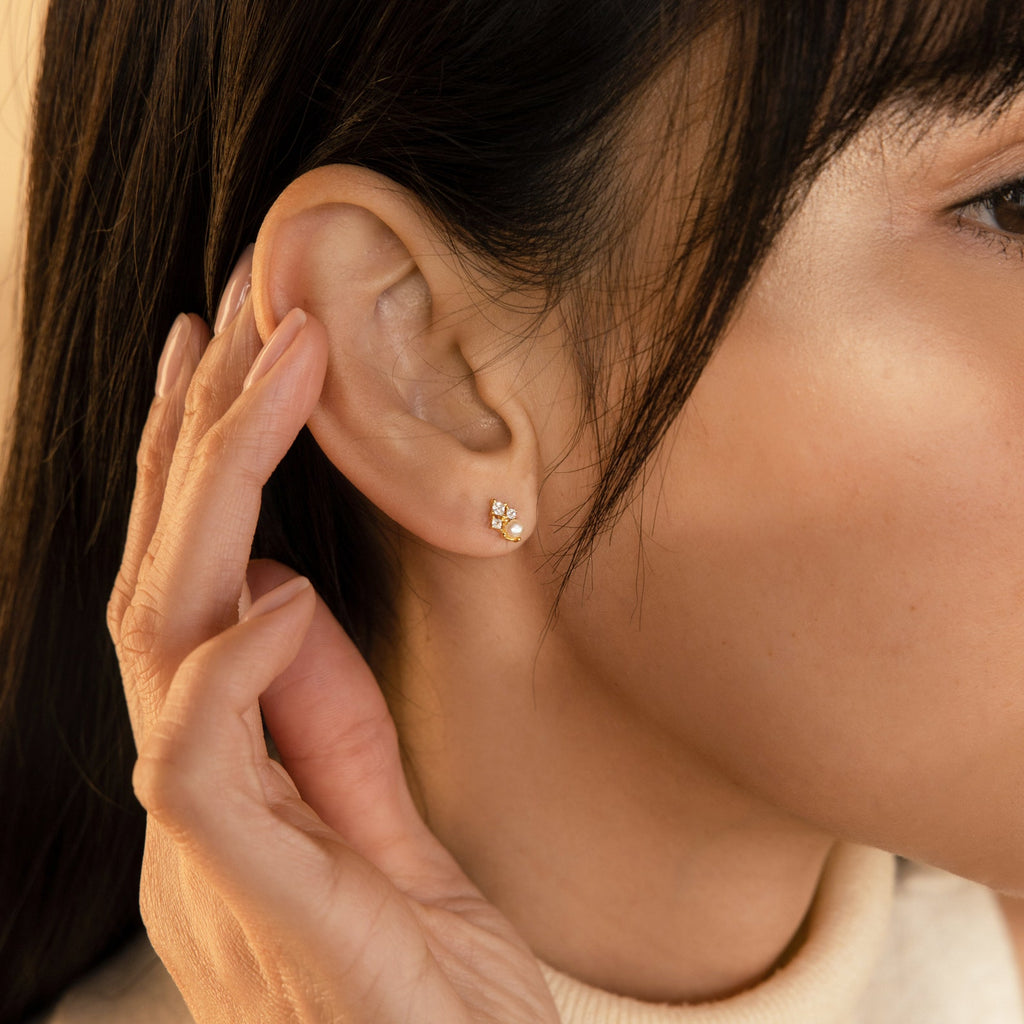 A person touches their ear, wearing the Angelina Pearl Studs—elegant gold earrings adorned with three tiny pearls.