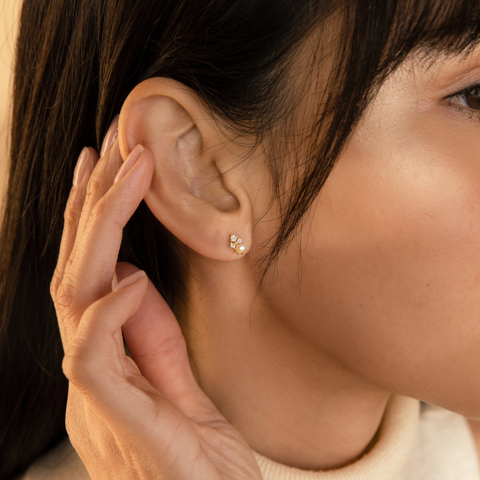 A person touches their ear, wearing the Angelina Pearl Studs—elegant gold earrings adorned with three tiny pearls.