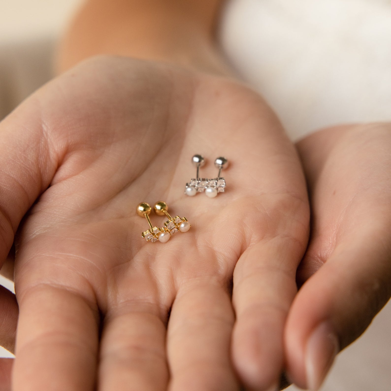 A hand holds the Angelina Pearl Studs—elegant earrings featuring timeless pearls set in gold and silver designs with gemstone details.