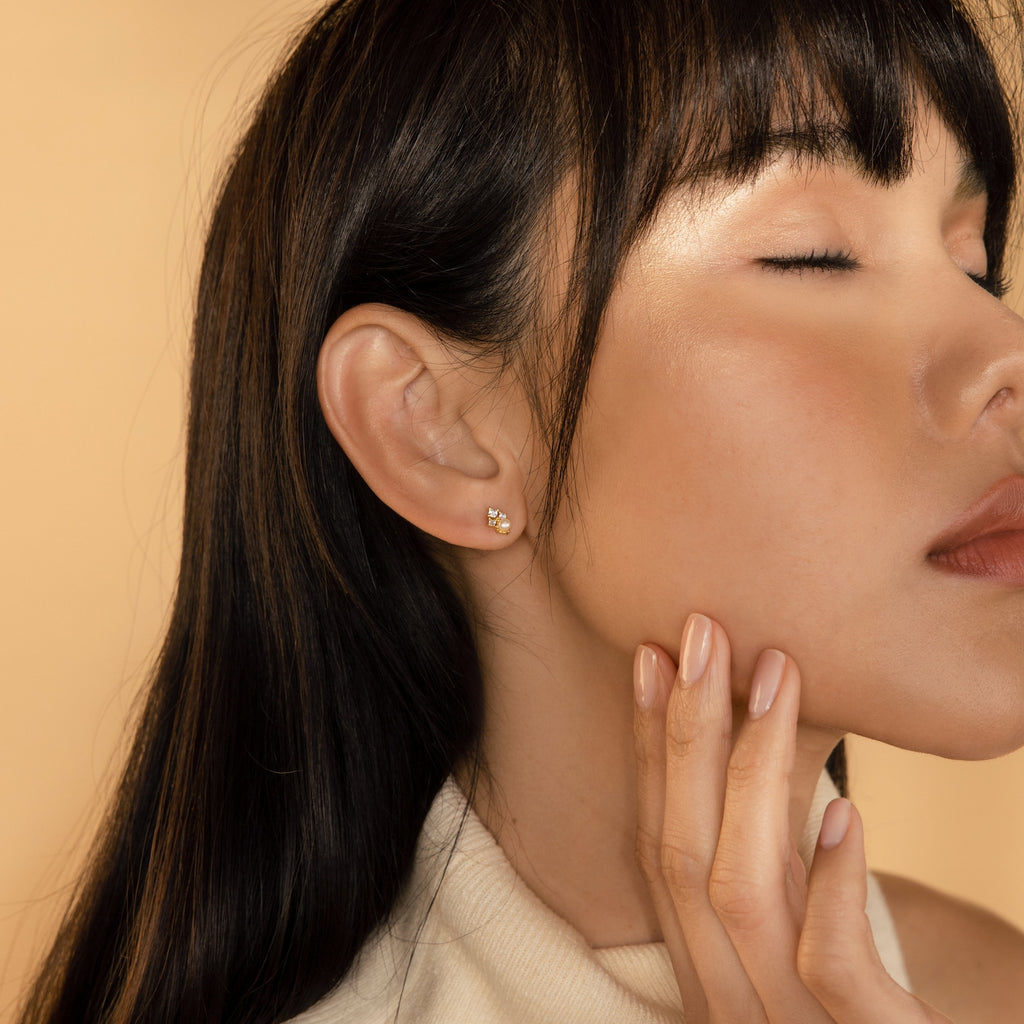 A woman with closed eyes gently touches her face, showcasing the elegant Angelina Pearl Studs against a beige background.