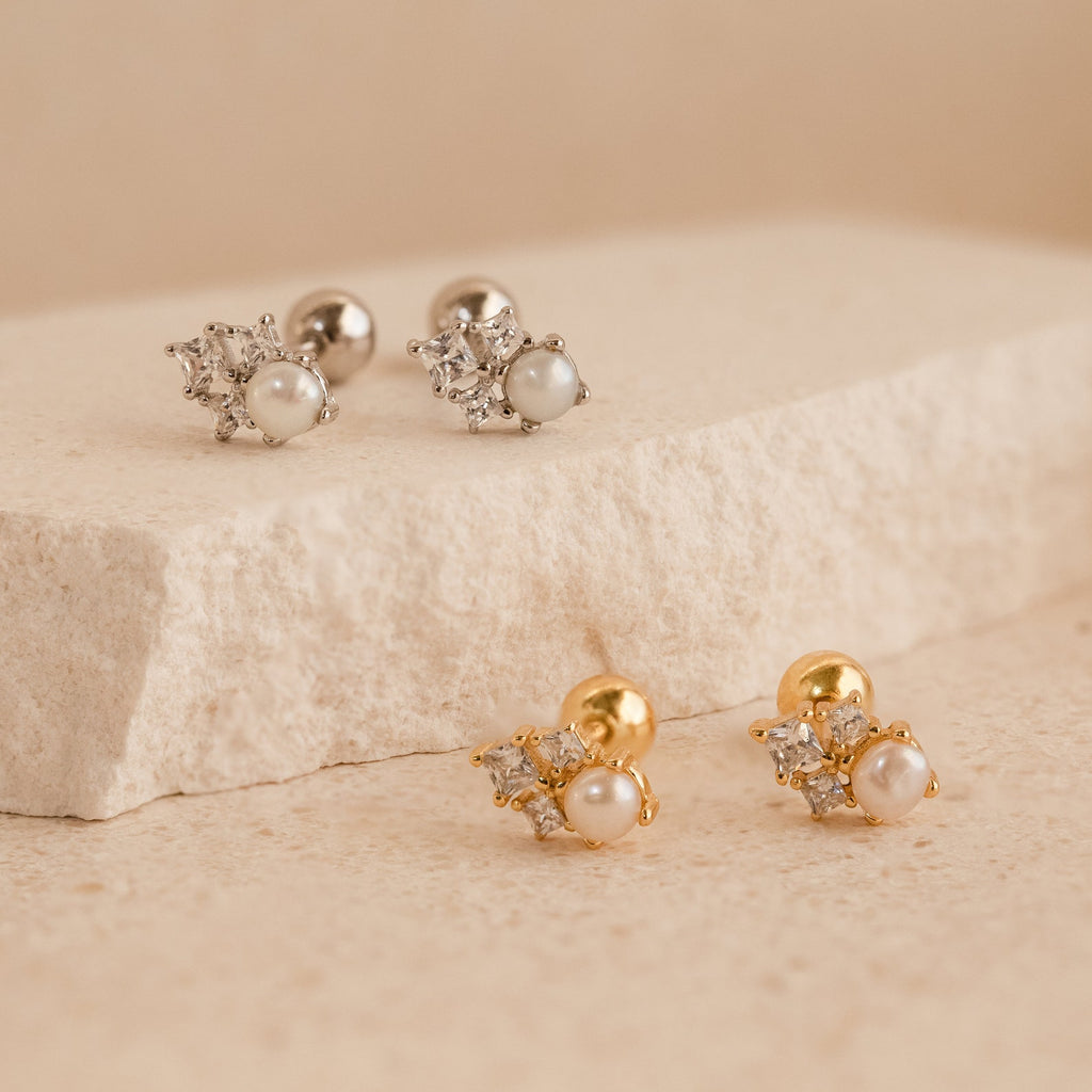 Two pairs of Angelina Pearl Studs, each featuring a pearl design and sparkling crystals—one in silver, one in gold—displayed on a stone surface.