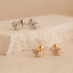 Two pairs of Angelina Pearl Studs, each featuring a pearl design and sparkling crystals—one in silver, one in gold—displayed on a stone surface.
