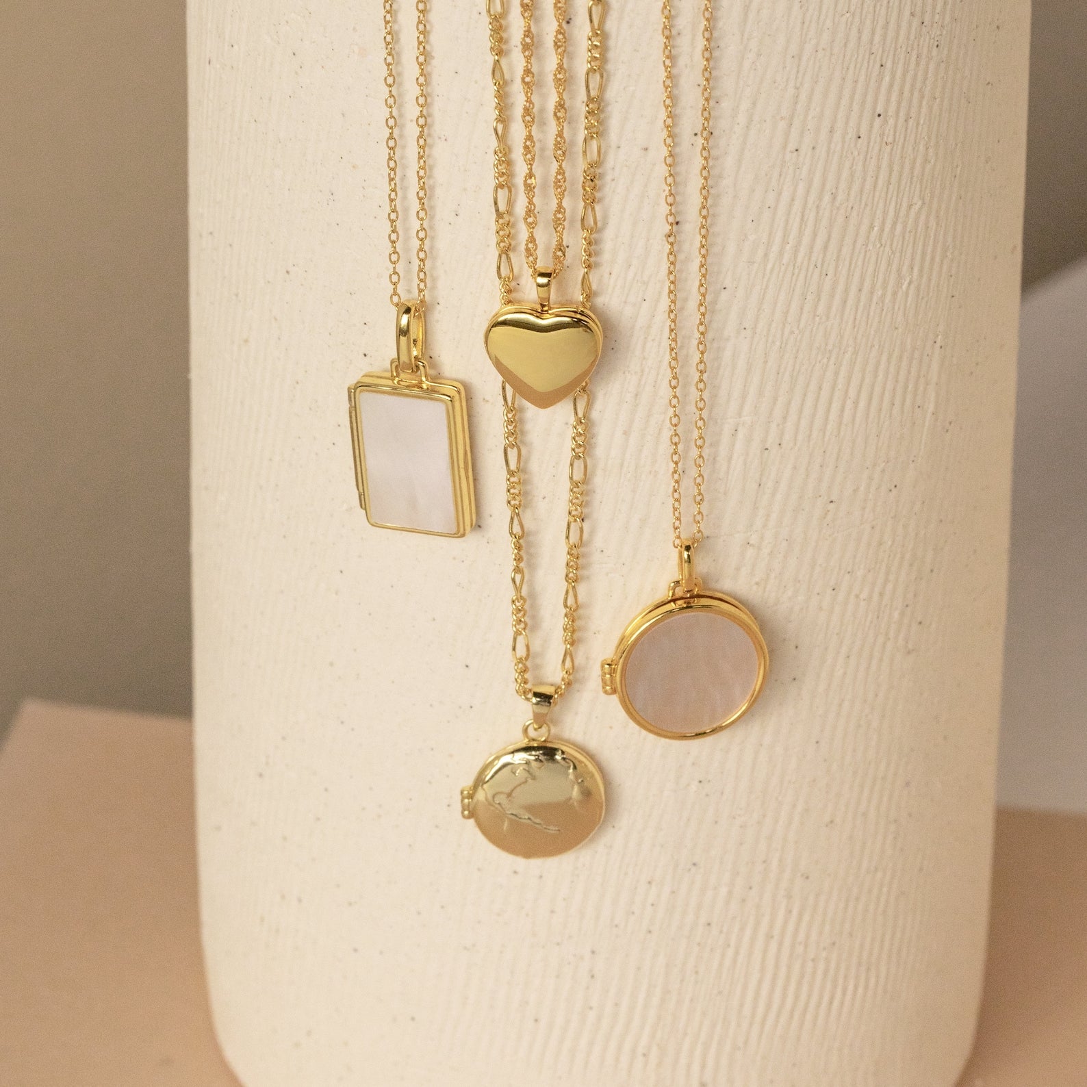 The Pearl Locket Necklace, featuring a Mother of Pearl accent and vintage-inspired pendant, is displayed among four gold necklaces on a textured white cylindrical stand.