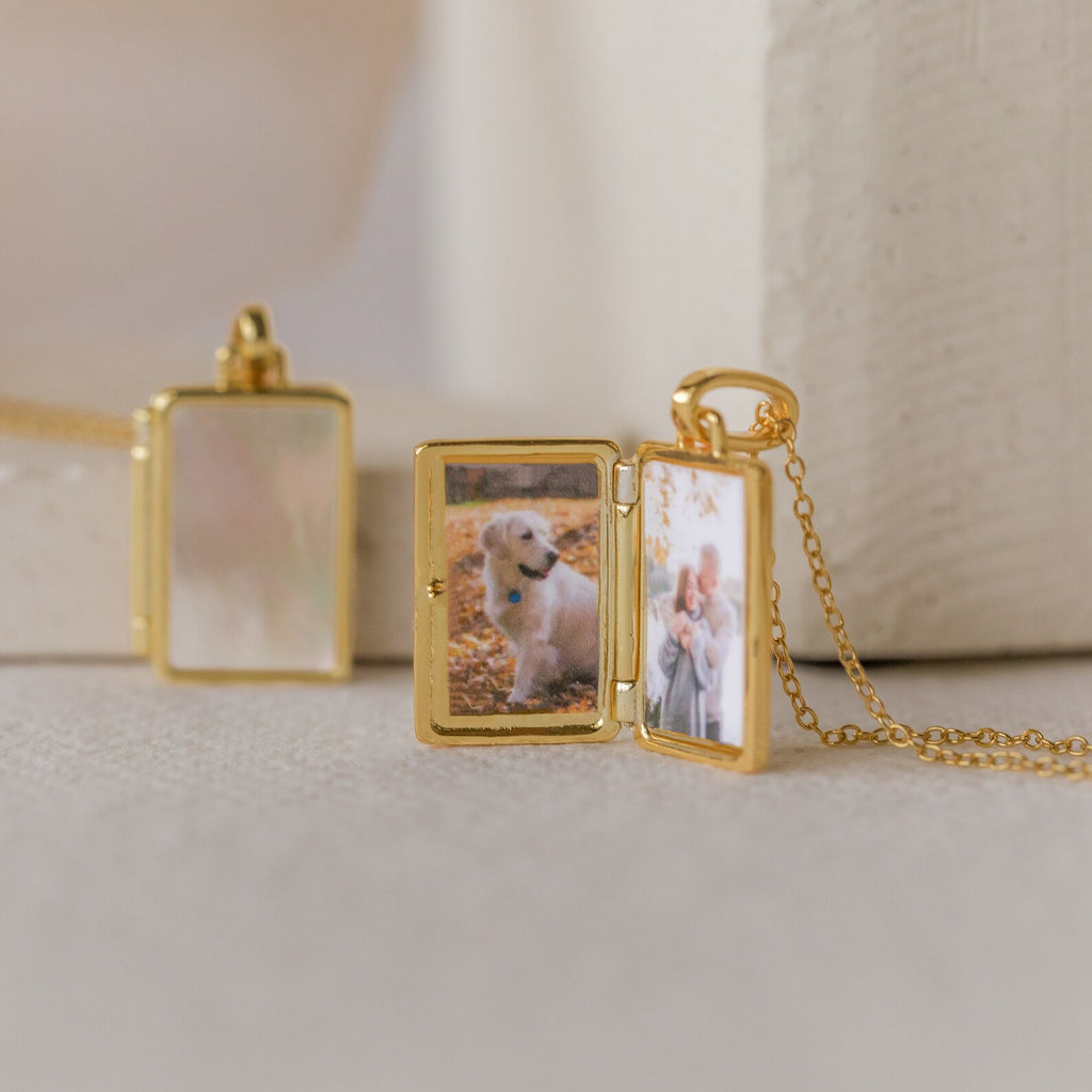 A Gold vintage locket, similar to the Pearl Locket Necklace, rests on a light surface with photos of a dog and two people inside—a classic keepsake.