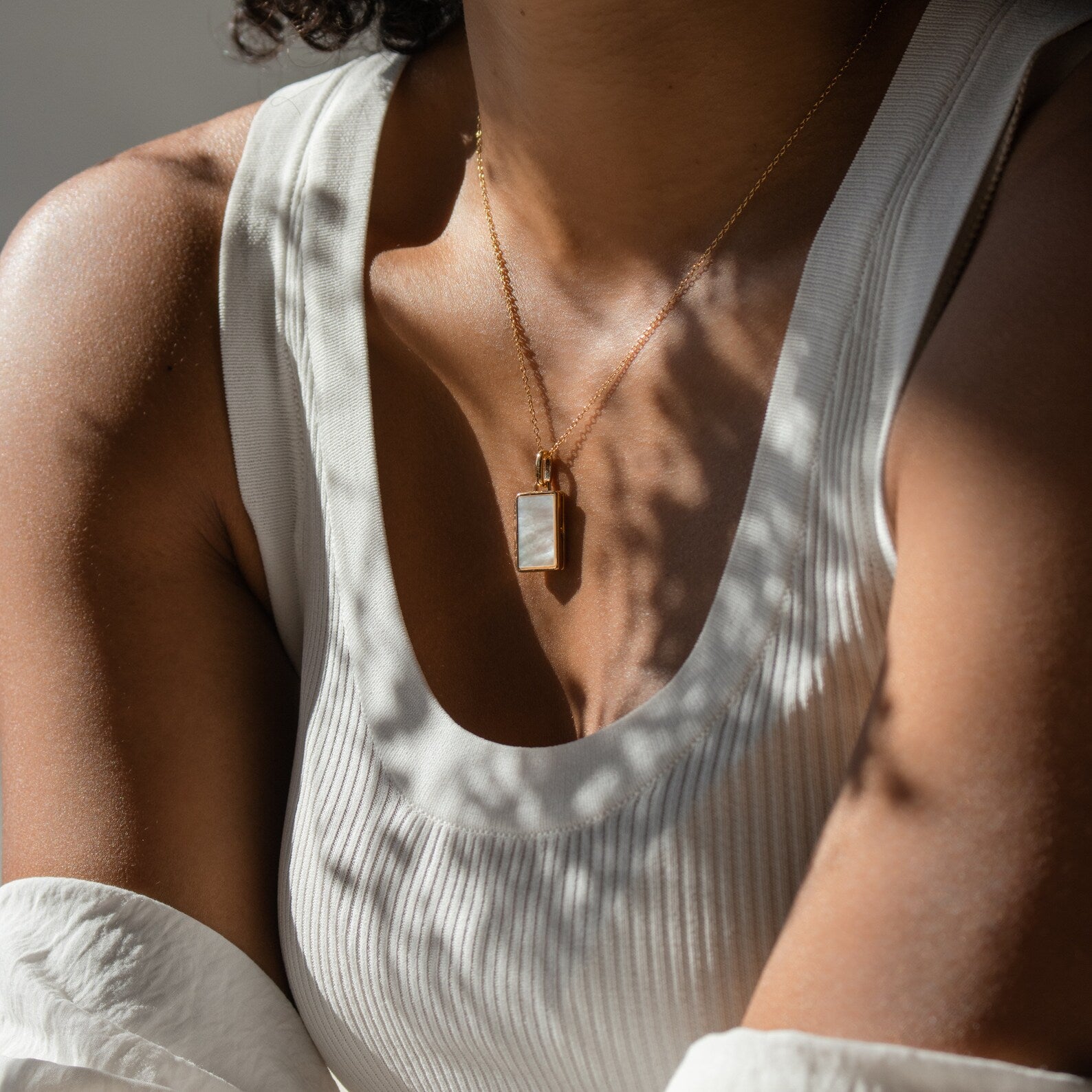 Wearing a white ribbed tank top, a person sits in sunlight with leafy shadows across their chest, showcasing the Endless Love Locket Necklace with its sterling silver rectangular pendant.