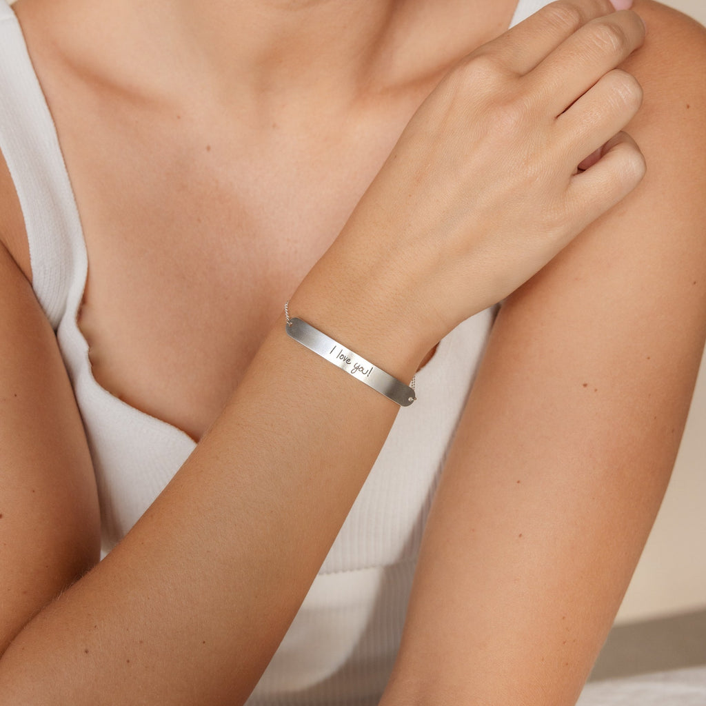 A woman wears the Slider Handwriting Bracelet, engraved with "I love you," as she gently rests her hand on her arm.