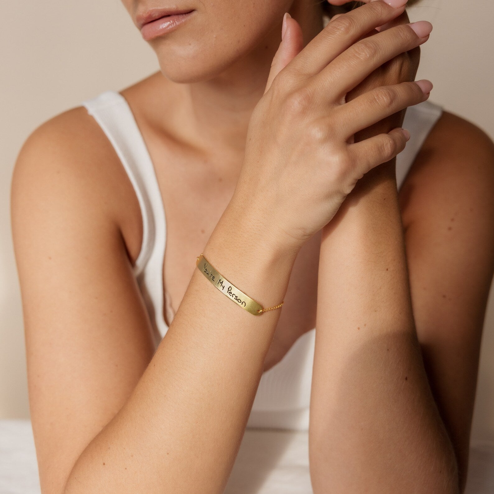 A woman in a white tank top wears the Slider Handwriting Bracelet, a dainty personalized piece featuring engraved text, with her hands clasped near her face.