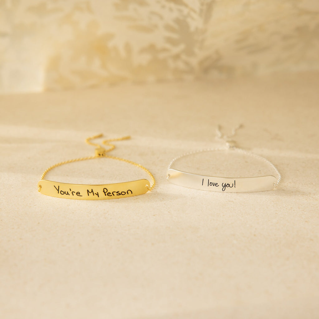 Two Slider Handwriting Bracelets—one gold engraved with "You’re My Person" and one silver with "I love you!"—rest on a beige surface, making ideal personalized keepsakes to cherish daily.