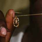A hand holds the Rainbow Initial Necklace, showcasing a gold R pendant encircled by colorful gems—a perfect personalized jewelry piece.