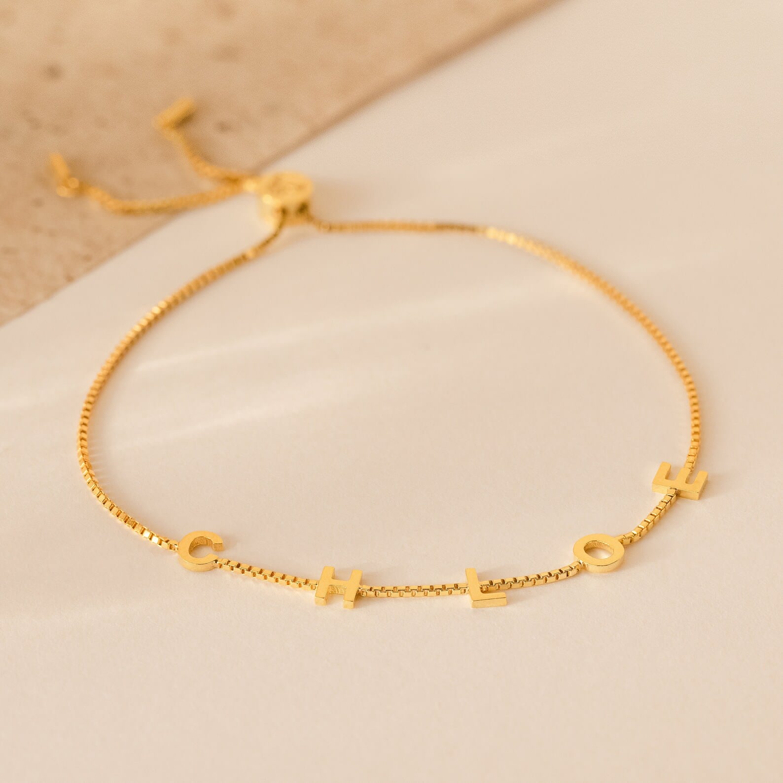 The Alex Slider Bracelet in Box Chain features gold dangling letters spelling CHLOE on a neutral background—an adjustable, personalized jewelry piece that makes a thoughtful customizable gift.