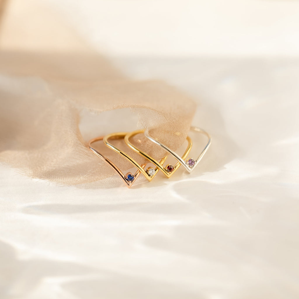 Three elegant Chevron Birthstone Rings, each set with sparkling gemstones, are displayed on soft beige fabric in gentle sunlight—a stunning showcase of birthstone jewelry.
