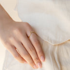 A hand with neatly manicured nails wears a delicate Chevron Birthstone Ring on the index finger, set against a cream dress.
