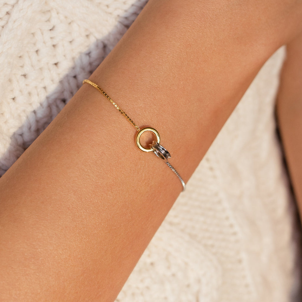 The Interlocking Circles Bracelet, featuring minimalist gold and silver rings, is shown on a wrist against cream knit fabric—an elegant charm bracelet ideal as an anniversary gift.