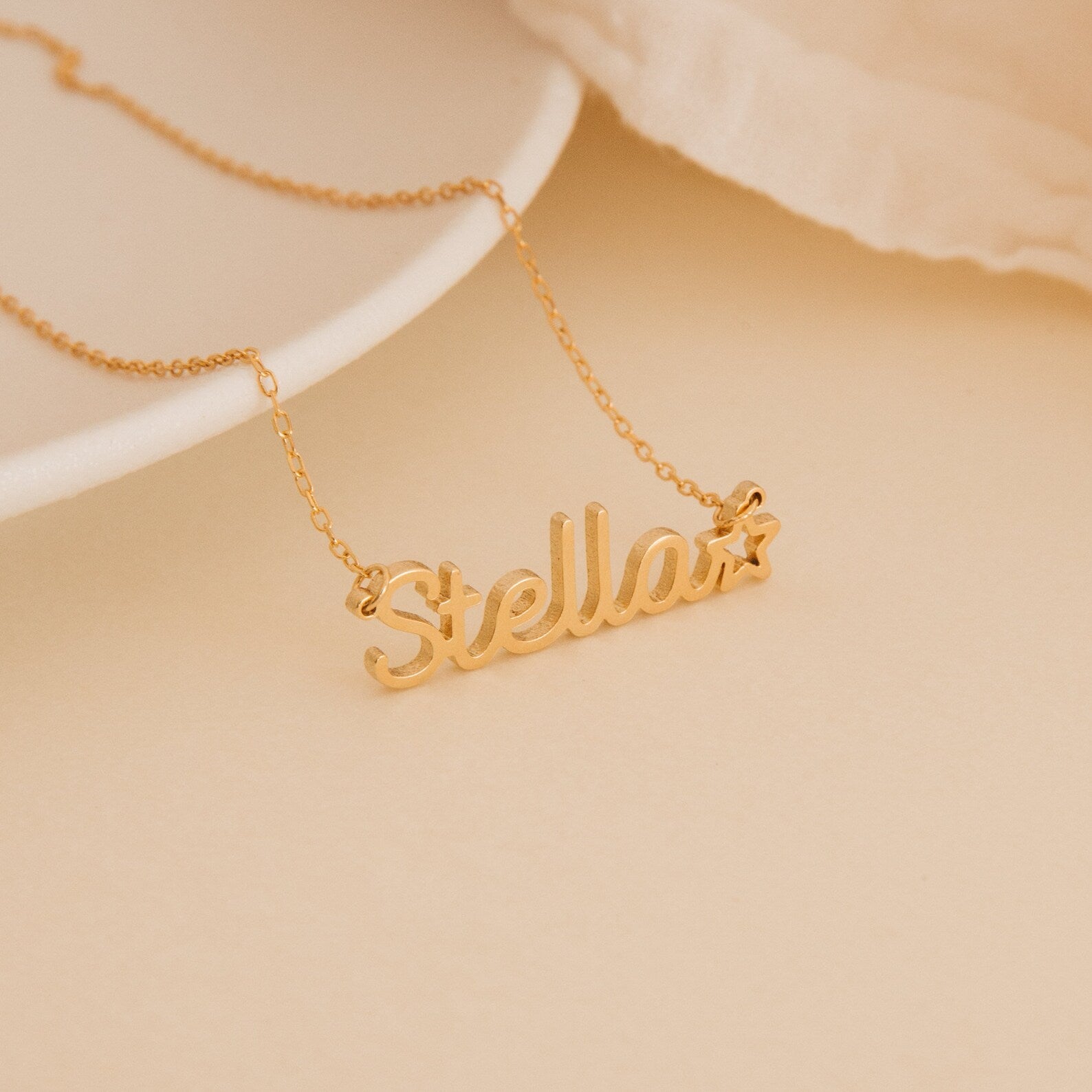 The Maine Kid's Name Necklace, customized with the name Stella and a small star charm at the end, is shown on a beige surface.