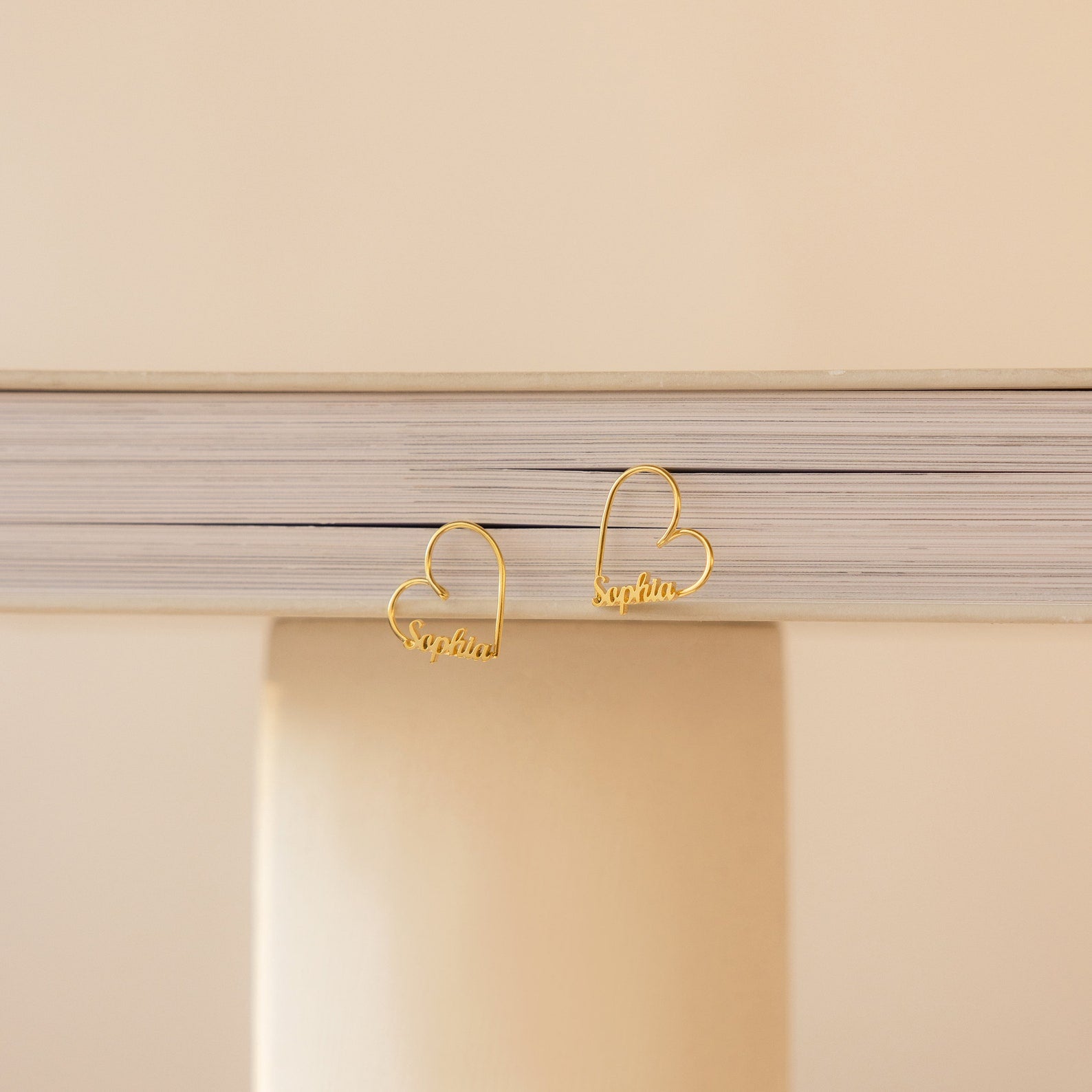 The London Heart Name Earrings, gold heart-shaped hoops featuring the name Sophia, are displayed on a stack of books against a beige background—an elegant personalized gift.