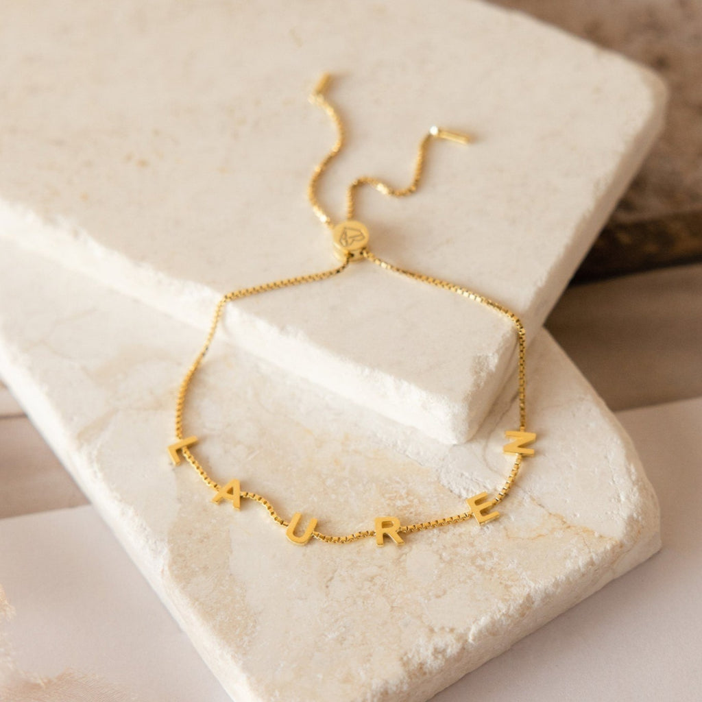 The Alex Slider Bracelet in Box Chain features adjustable length and customizable letter charms—shown here spelling LAUREN on stone slabs—making it a perfect personalized jewelry gift.