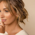 Woman with wavy hair wearing minimalist Large Ribbed Link Hoops and a cream sweater, smiling softly against a beige background.