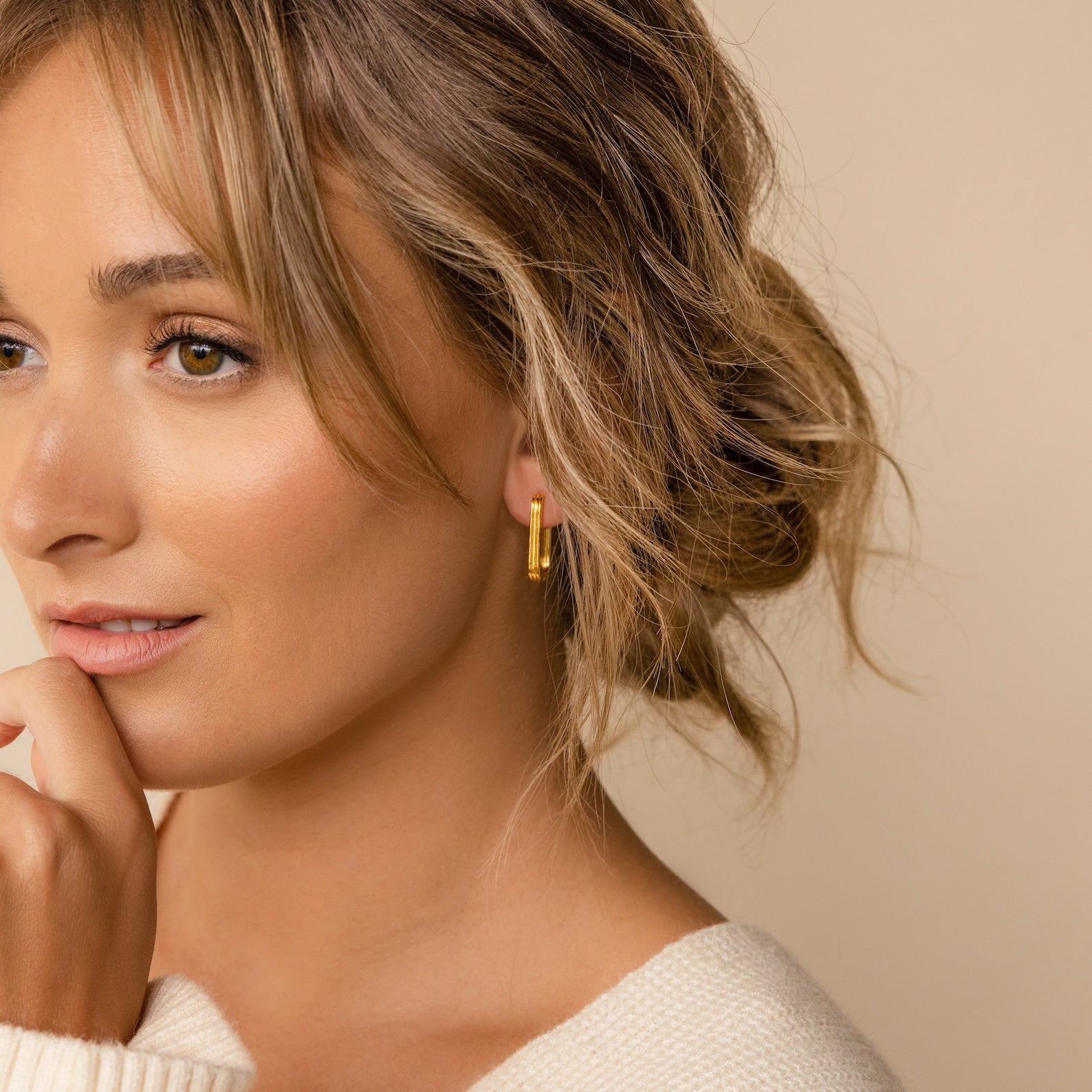 Woman with wavy hair wearing minimalist Large Ribbed Link Hoops and a cream sweater, smiling softly against a beige background.