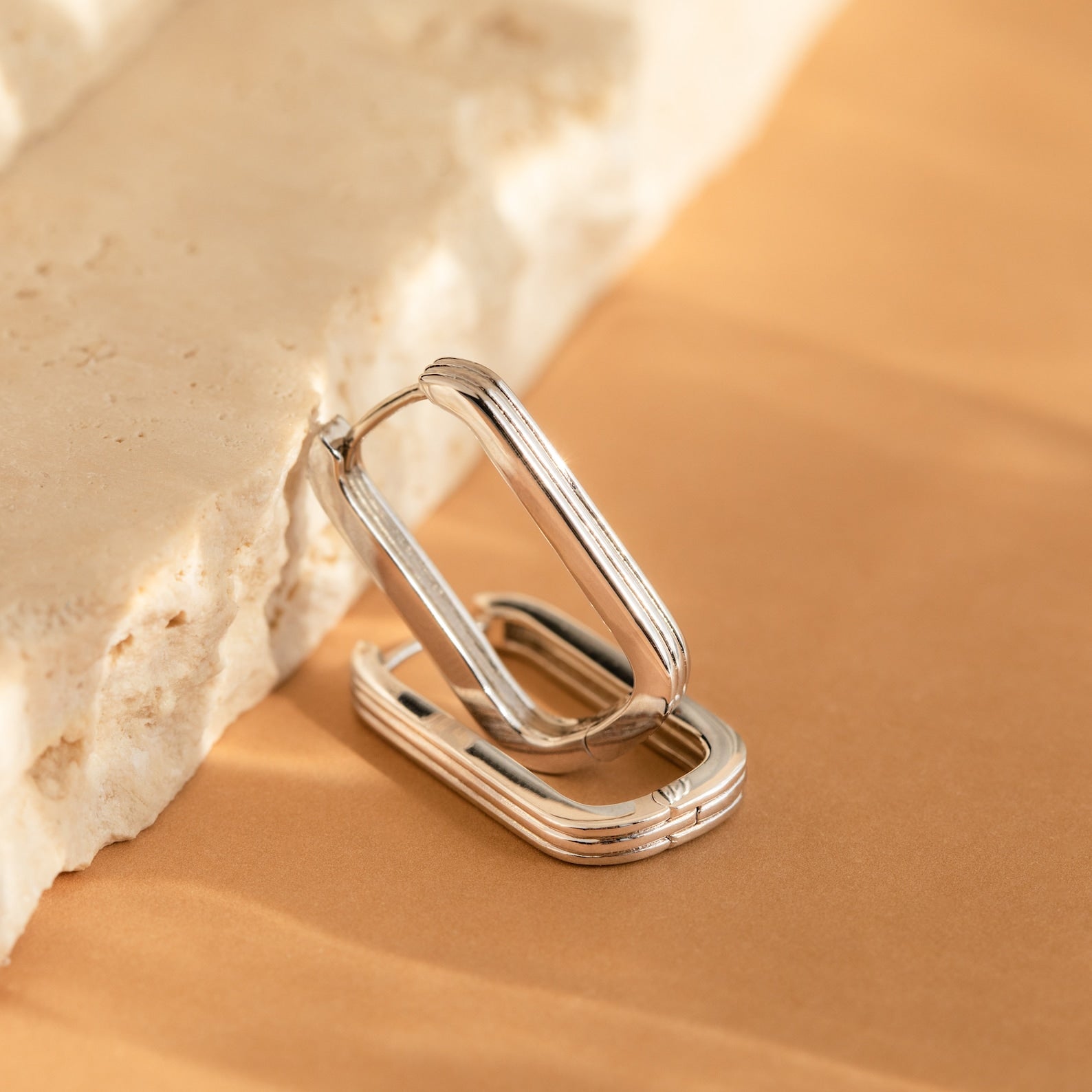 Large Ribbed Link Hoops in silver, featuring a rectangular ribbed design, rest against a textured beige stone on a tan surface, exemplifying minimalist jewelry style.
