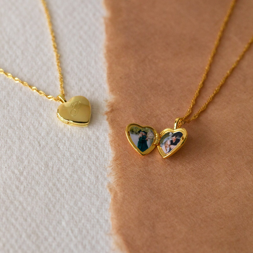 The Initial Heart Locket Necklace features two gold heart lockets on chains—one closed, one open displaying small photos—resting on textured paper, making it a timeless piece ideal for adding your engraved initial.