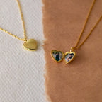 The Initial Heart Locket Necklace features two gold heart lockets on chains—one closed, one open displaying small photos—resting on textured paper, making it a timeless piece ideal for adding your engraved initial.