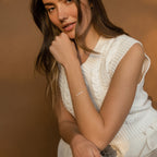 A woman in a white knitted sleeveless top poses against a brown backdrop, resting her chin on her hand and showcasing the Venice Tiny Name Bracelet for a touch of elegant, minimal jewelry.