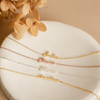 The Venice Tiny Name Bracelet, a delicate minimal jewelry piece, is displayed on a round white surface with dried flowers.