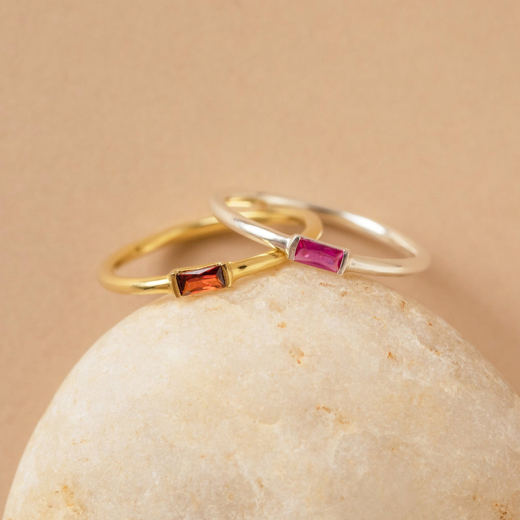 Two delicate Baguette Birthstone Rings with rectangular gemstones—one gold with orange, one silver with pink—rest on a smooth rock. Personalize this ring for a unique way to celebrate your story or as a thoughtful birthstone gift.