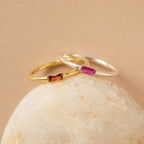 Two delicate Baguette Birthstone Rings with rectangular gemstones—one gold with orange, one silver with pink—rest on a smooth rock. Personalize this ring for a unique way to celebrate your story or as a thoughtful birthstone gift.