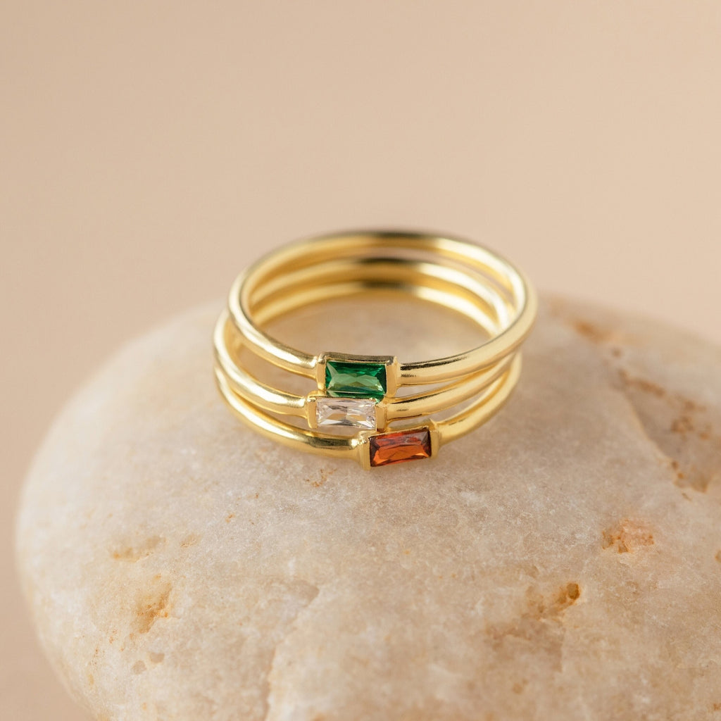 Three gold Baguette Birthstone Rings with rectangular green, clear, and brown stones are elegantly stacked on a smooth beige stone—ideal for a personalized touch.