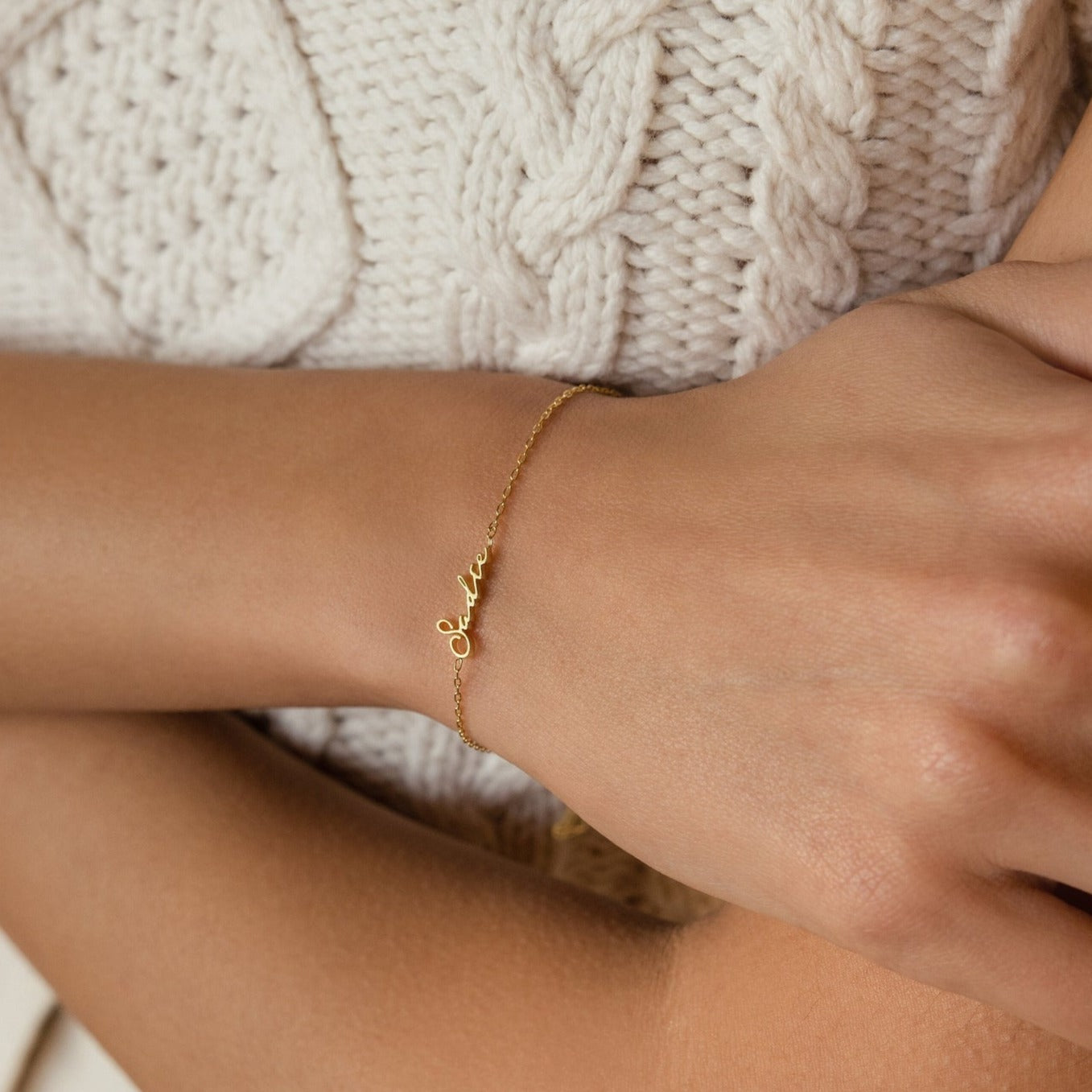 The Venice Tiny Name Bracelet, a minimal jewelry piece with a cursive gold name, adds an elegant touch when paired over a cream knit sweater.