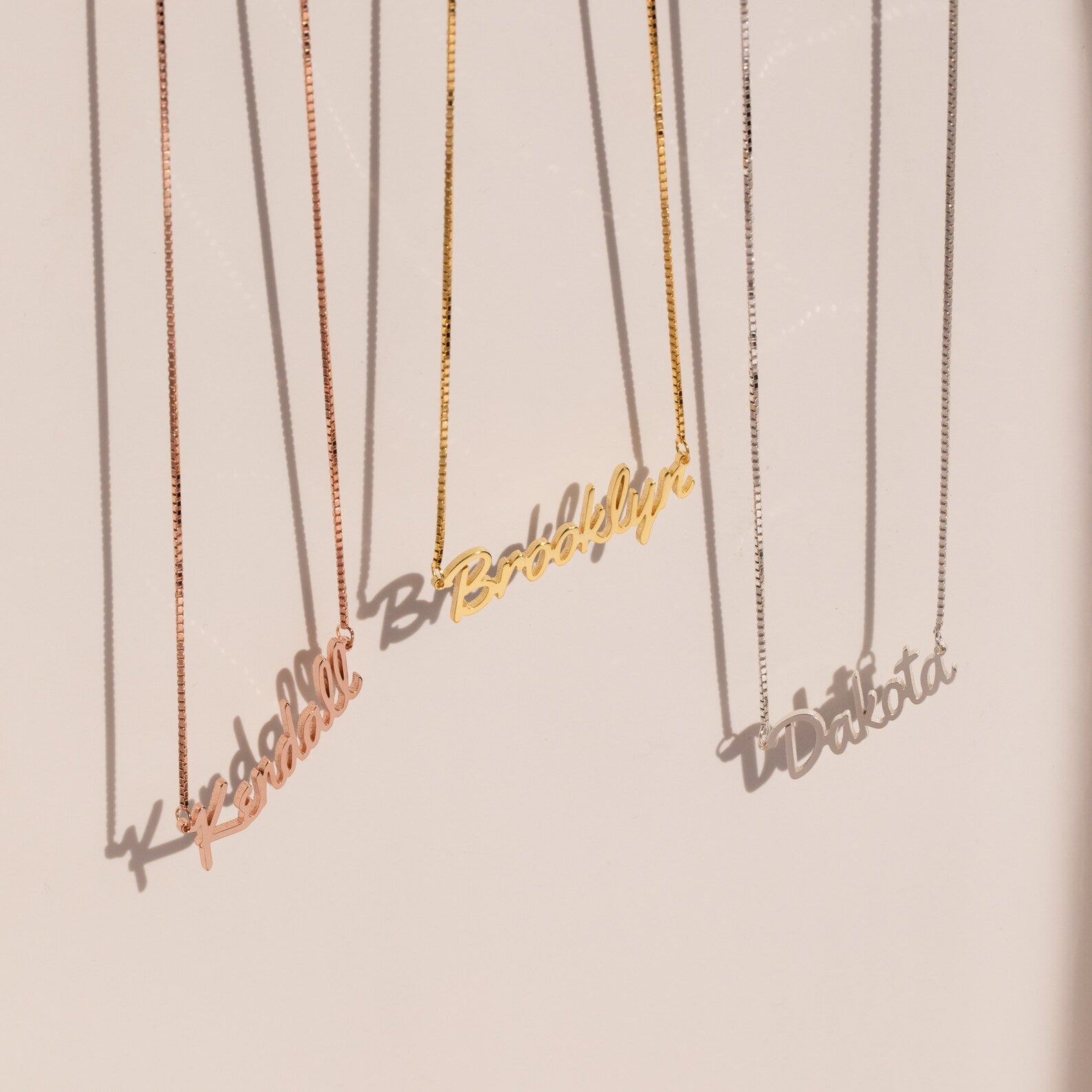 Three Casella Name Necklaces in box chains, each personalized with Kendall, Brooklyn, and Dakota in a dainty script font, hang elegantly—perfect as a custom gift.