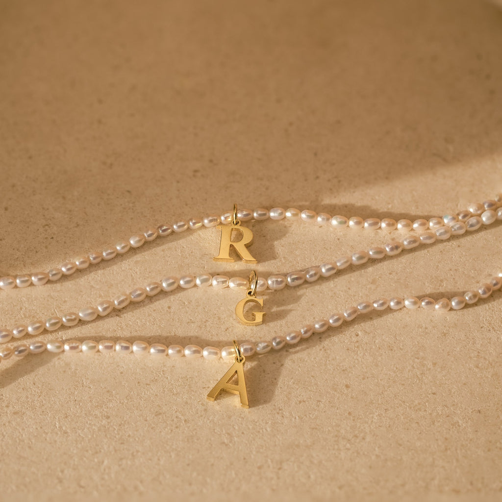 Three Newsletter Pearl Initial Necklaces with gold letter pendants R, G, and A rest on a beige surface—minimalist jewelry featuring customizable letter charms for a personal touch.