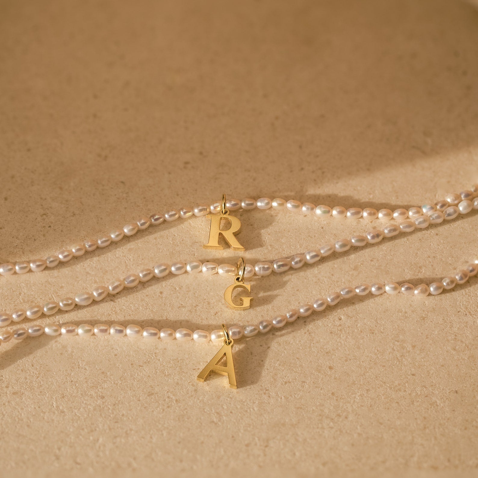 Three Newsletter Pearl Initial Necklaces with gold letter pendants R, G, and A rest on a beige surface—minimalist jewelry featuring customizable letter charms for a personal touch.