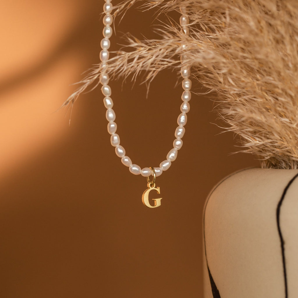 Minimalist jewelry at its best: the Newsletter Pearl Initial Necklace features a customizable initial charm and a gold G pendant, elegantly displayed beside dried pampas grass in a beige vase.