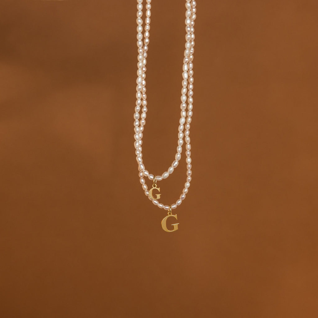 The Newsletter Pearl Initial Necklace in Rose Gold, featuring a customizable initial charm, hangs against a plain brown background—an ideal choice for fans of minimalist jewelry.