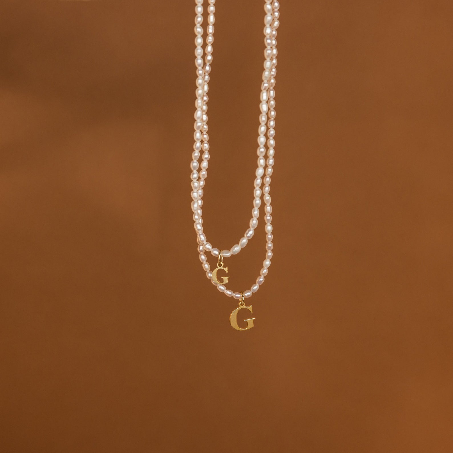 The Newsletter Pearl Initial Necklace in Rose Gold, featuring a customizable initial charm, hangs against a plain brown background—an ideal choice for fans of minimalist jewelry.
