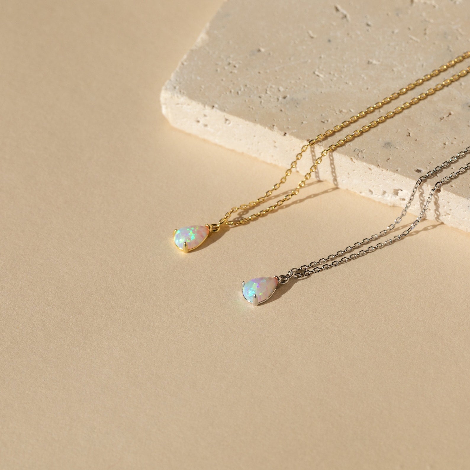 Two Tabitha Opal Drop Necklaces, each with a delicate teardrop charm—one in gold, one in silver—are displayed on a beige stone background. A romantic gift perfect for any occasion.