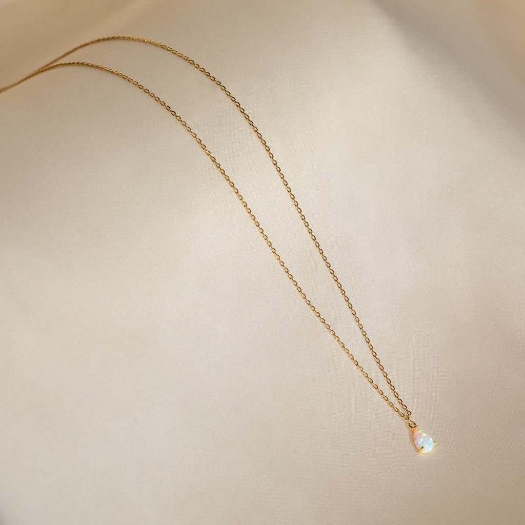 The Tabitha Opal Drop Necklace features a delicate opal teardrop charm on a gold chain, displayed on soft beige fabric—perfect as a romantic gift.