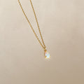 A gold chain necklace featuring a small, iridescent teardrop charm pendant—the Tabitha Opal Drop Necklace—displayed on a light beige background, perfect as a romantic gift.