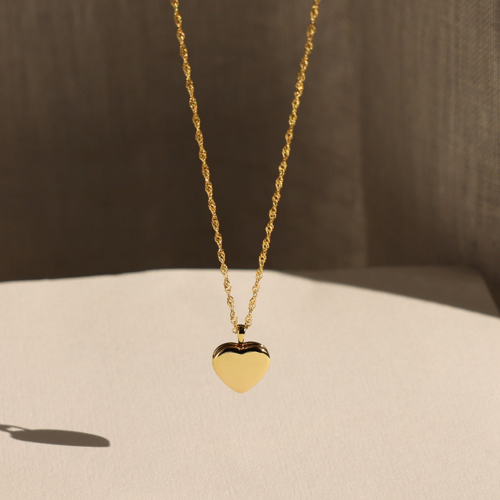 The Initial Heart Locket Necklace, a gold heart-shaped pendant with an engraved initial, is displayed against a soft neutral background.
