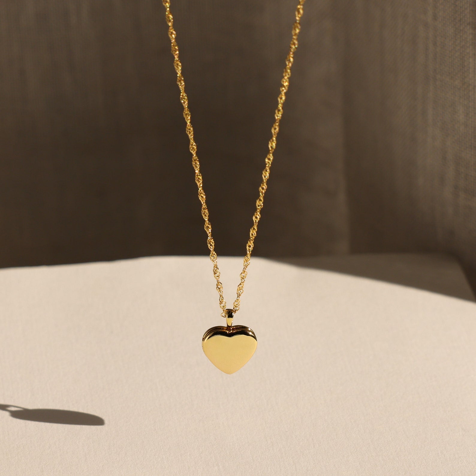 The Initial Heart Locket Necklace, a gold heart-shaped pendant with an engraved initial, is displayed against a soft neutral background.